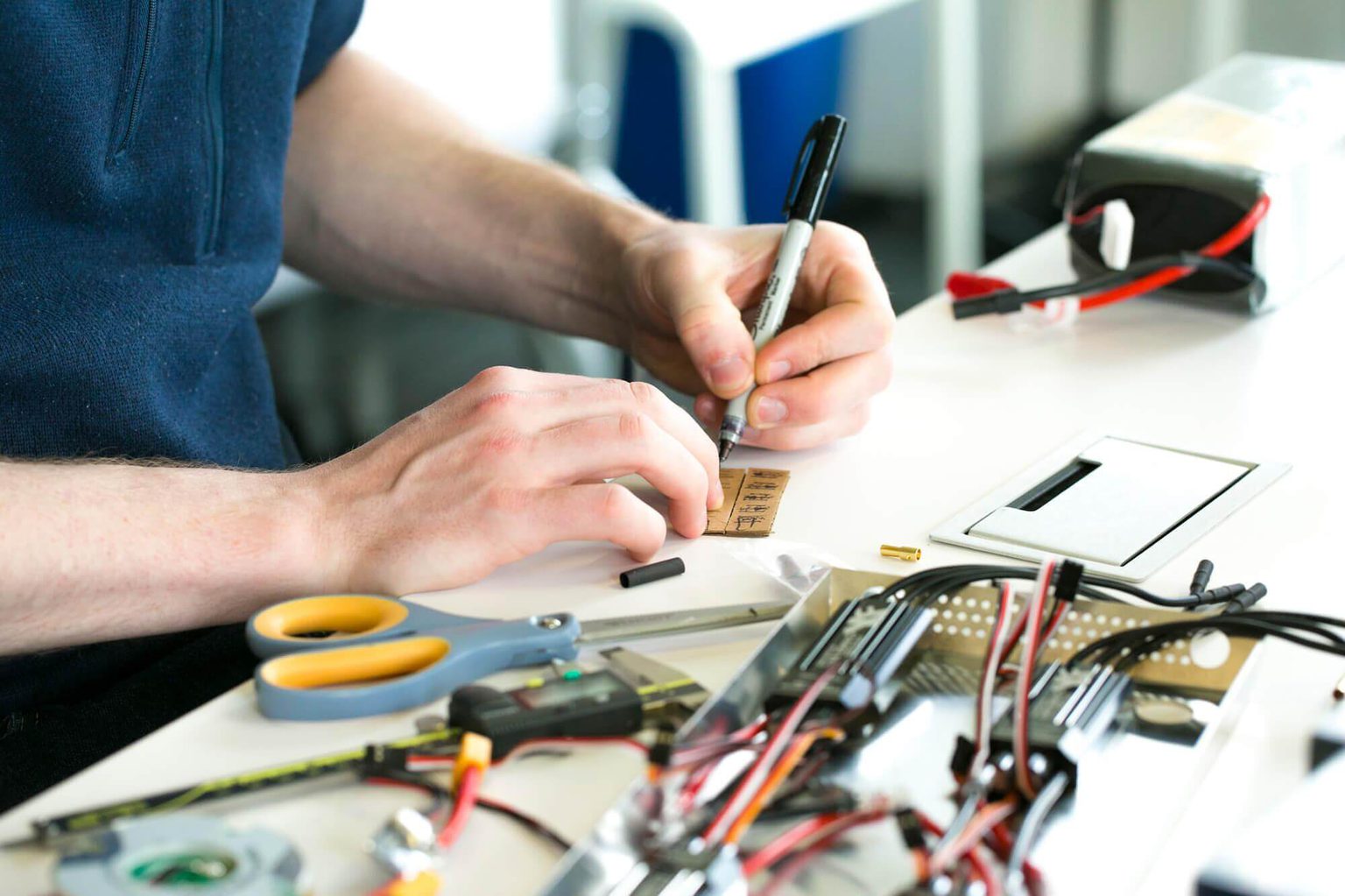 Man in blue shirt at work station soldering with electrical components scattered across table