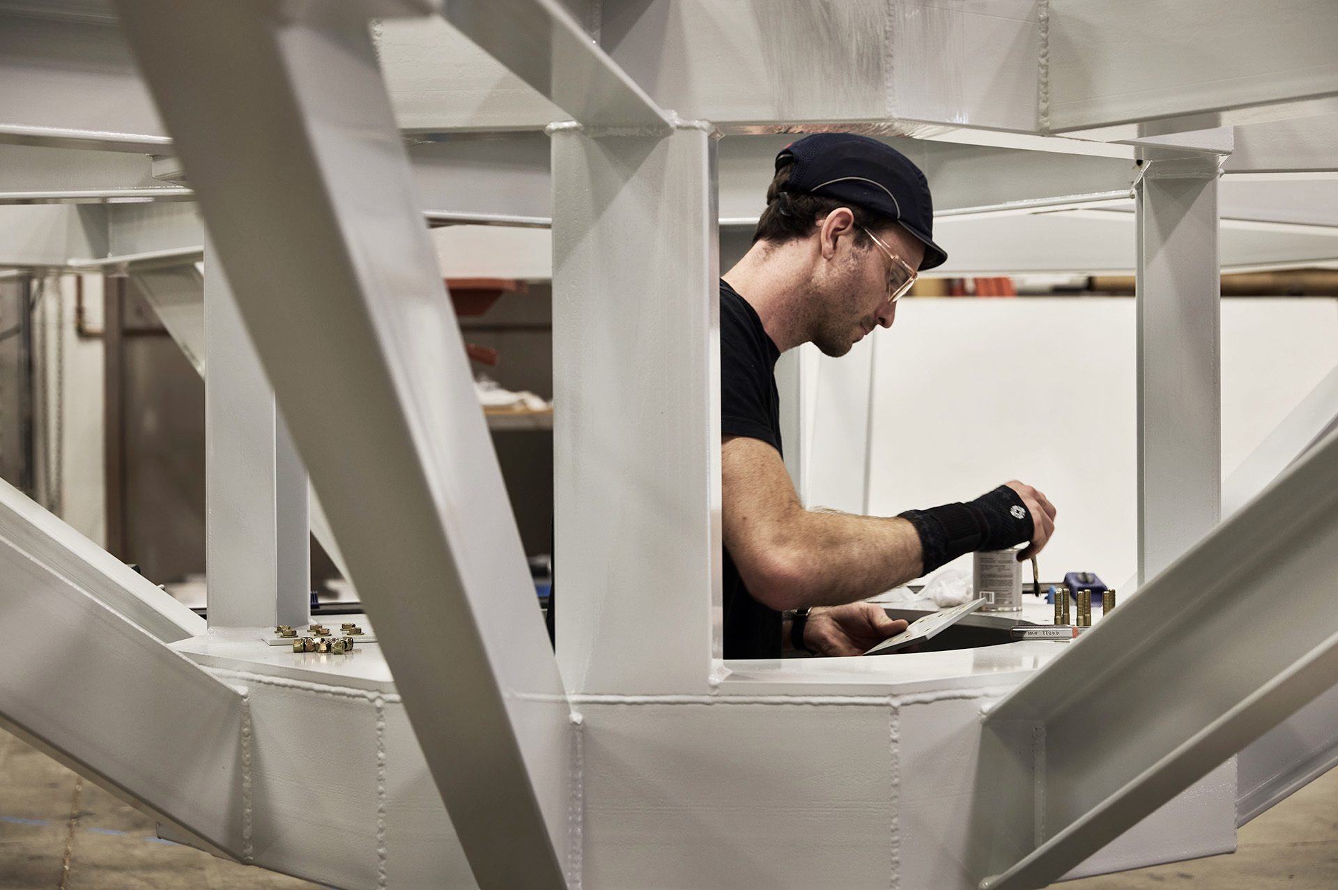 Man in black shirt and black baseball hat standing inside a large metal structure with screw driver in hand