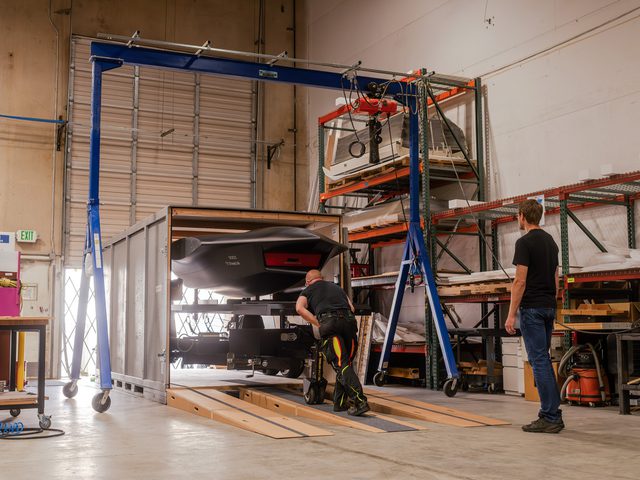 Man in black watching another man push load an aircraft into a shipping crate