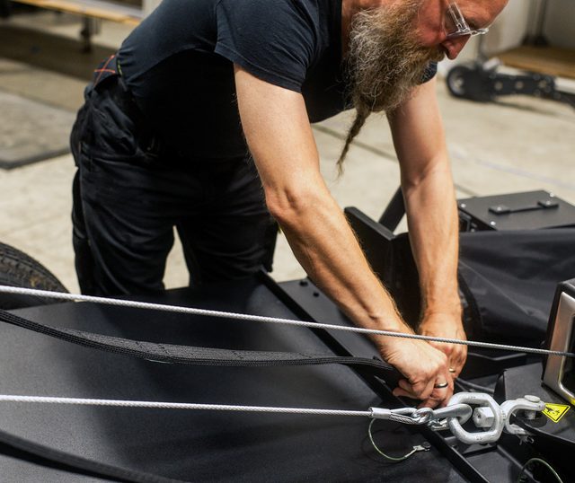 Man with long braided beard dressed in black bending over a model trailer attaching a wire with a clasp
