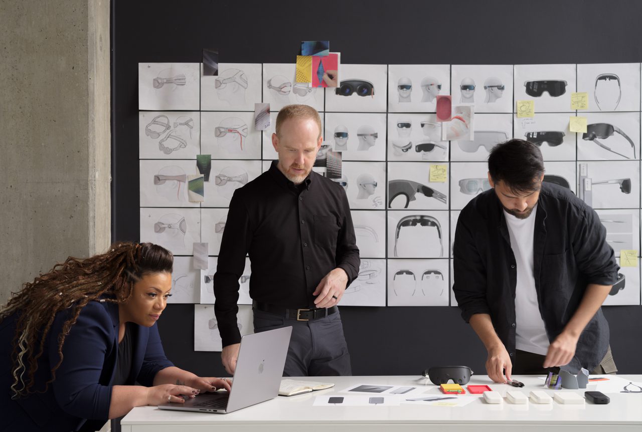 Team of designers looking at 3D printed prototypes with wall of colorful sketches behind them