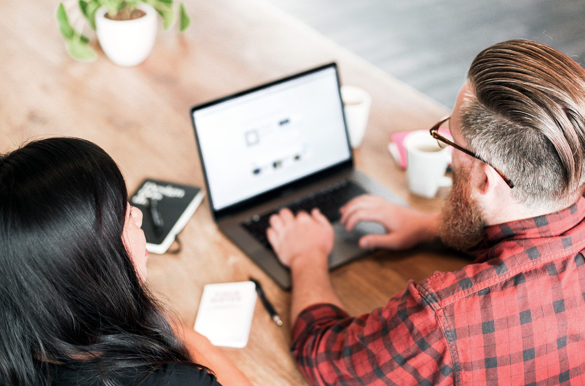 Man in red plaid shirt seated with women with dark hair looking at a laptop